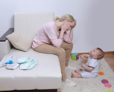A stressed mother sitting on a couch while their child is sitting on the ground staring at the mother