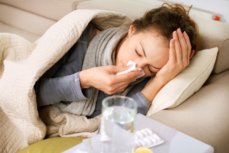 Sick woman lying on couch with tissue, headache, and medication nearby