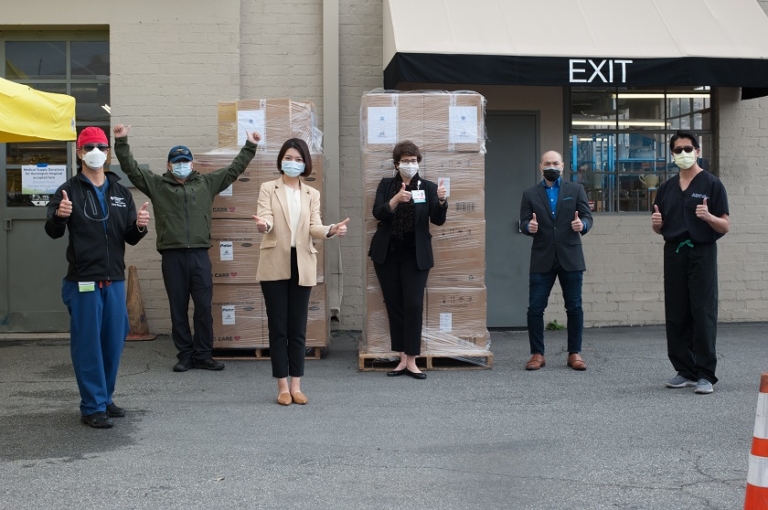 Six masked individuals standing beside donated medical supply pallets outdoors